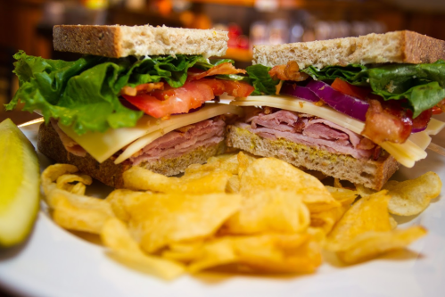 A close-up of a sandwich with layers of lettuce, tomato, cheese, ham, and bacon, served with potato chips and a pickle.