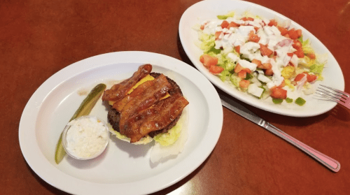A plate with a bacon cheeseburger, pickle, and a side of salad with tomatoes and cucumbers.