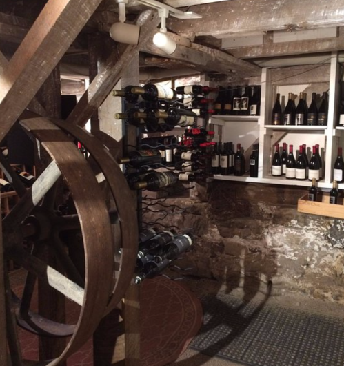 A rustic wine cellar featuring wooden beams, a wine rack with bottles, and shelves lined with various wines.