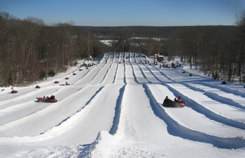 A snowy landscape with multiple tubing lanes, people sliding down on inflatable tubes under a clear blue sky.