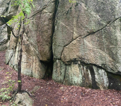 A large rock formation with cracks, surrounded by earthy ground and sparse vegetation.
