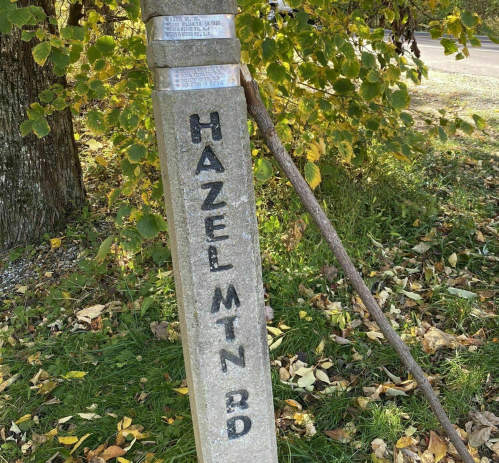 A concrete post with "HAZEL MTN RD" engraved, surrounded by grass and autumn leaves.