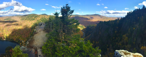 A panoramic view of colorful autumn foliage and mountains under a blue sky with scattered clouds.
