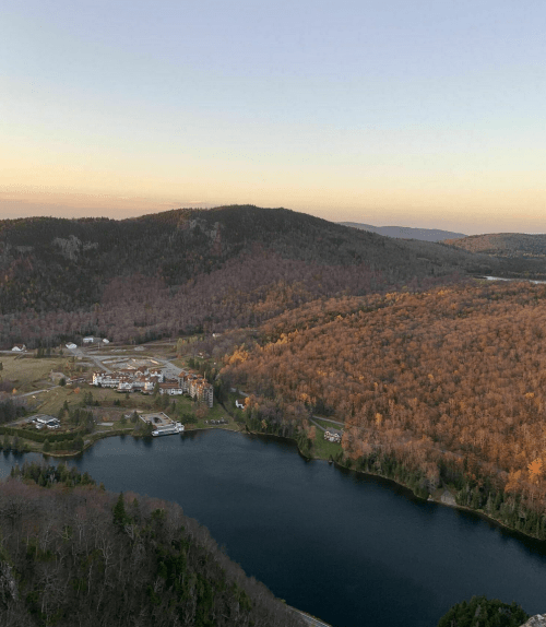 Aerial view of a serene lake surrounded by wooded hills and a small village at sunset.