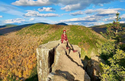 A person sits on a rocky ledge overlooking a colorful autumn landscape with mountains and a blue sky.