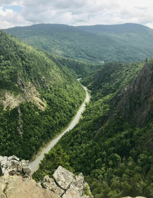 A winding road cuts through a lush green valley surrounded by steep, rocky hills under a cloudy sky.
