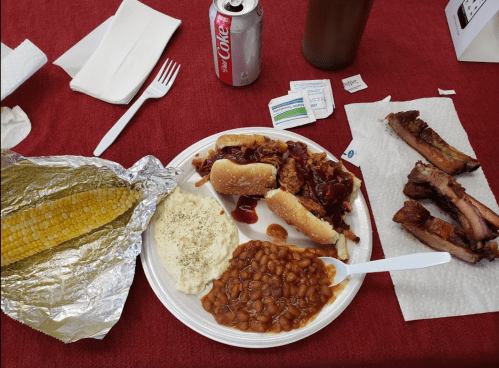 A plate with pulled pork sandwiches, baked beans, mashed potatoes, corn on the cob, and ribs, with a soda nearby.