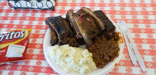 A plate of barbecue ribs, baked beans, potato salad, and a bag of Fritos on a checkered tablecloth.