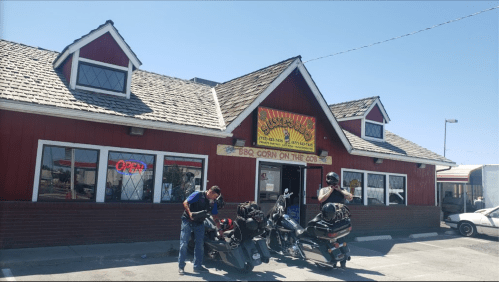 Two motorcyclists stand by their bikes in front of a red and white BBQ restaurant with a sign that says "Open."