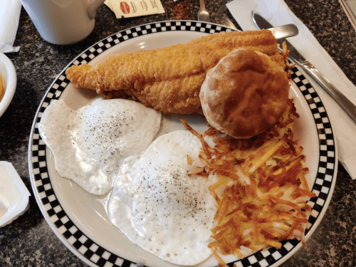 A plate with fried fish, two sunny-side-up eggs, hash browns, and a biscuit on a checkered rim.