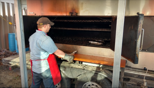 A person in a blue shirt and red apron is grilling food in a large outdoor smoker.