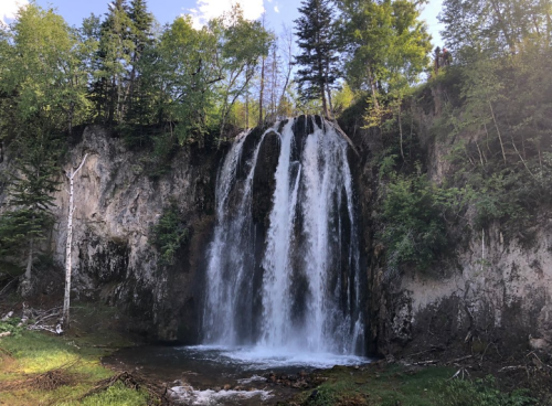 A cascading waterfall surrounded by lush green trees and rocky terrain under a clear blue sky.