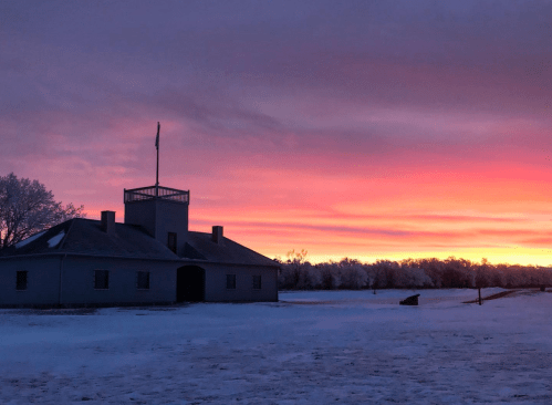 A historic building silhouetted against a vibrant sunset, with snow covering the ground and trees in the background.