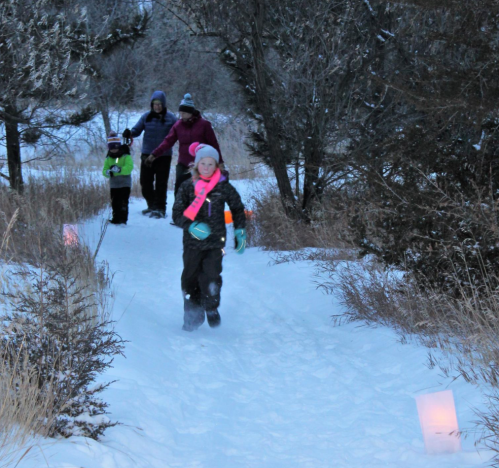 A group of people, including children, walking on a snowy path lined with lanterns in a winter landscape.
