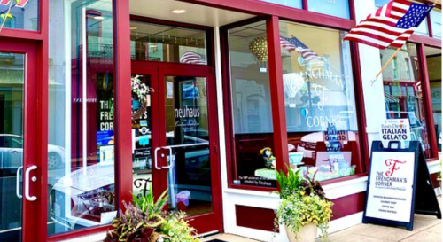 Exterior of a shop with large windows, flags, and a sign for Italian gelato, featuring colorful plants at the entrance.