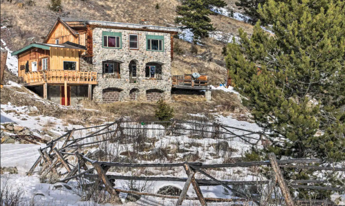 A rustic stone house with wooden accents, surrounded by snow and trees, set against a mountainous landscape.