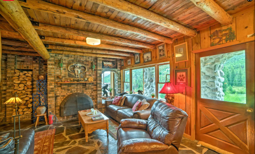 Cozy cabin interior with wooden beams, stone fireplace, leather sofas, and large windows overlooking nature.