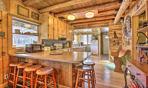 Cozy wooden kitchen with a bar, stools, and rustic decor, featuring large windows and natural light.