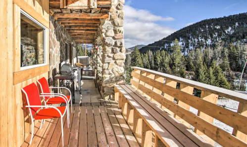 A wooden deck with red chairs overlooks a mountain landscape and trees under a blue sky.