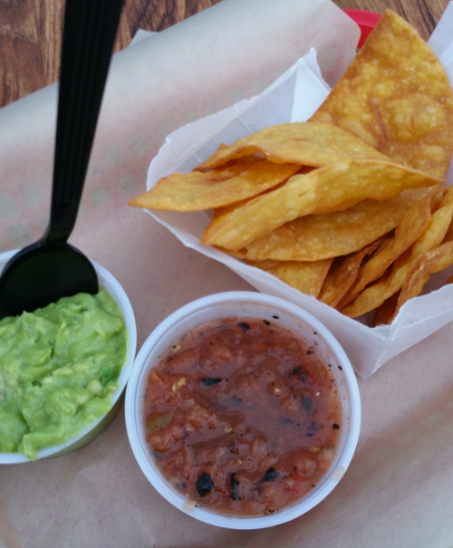 A serving of tortilla chips with guacamole and salsa in small containers on a paper-lined tray.