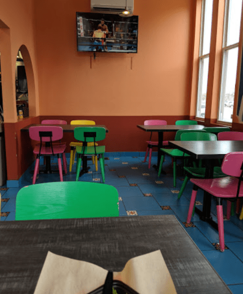 A colorful empty restaurant with pink and green chairs, blue tiled floor, and a TV showing a sports game.