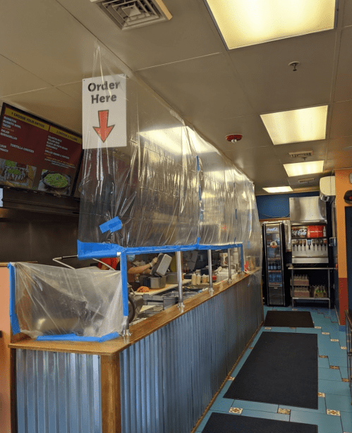 A restaurant counter with plastic barriers, a sign indicating "Order Here," and a drink station in the background.