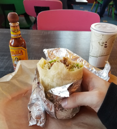 A hand holding a wrapped burrito with a bottle of hot sauce and a cup in the background on a colorful table.