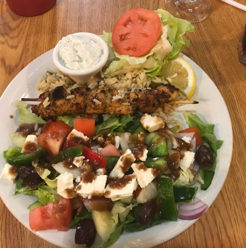 A plate with grilled chicken skewers, rice, a fresh salad with tomatoes, cucumbers, olives, and a side of sauce.