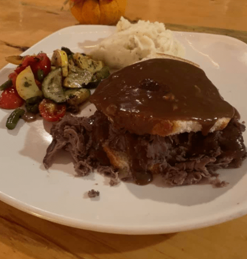 A plate featuring a roast beef sandwich with gravy, mashed potatoes, and a side of sautéed vegetables.