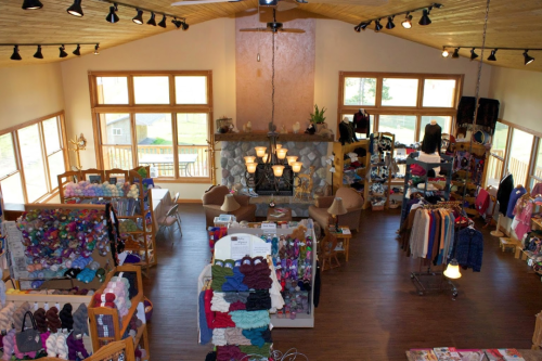 A cozy yarn shop with wooden decor, featuring shelves of colorful yarn, seating areas, and a stone fireplace.