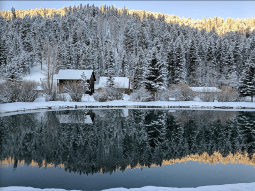 A serene winter scene with snow-covered trees reflecting in a calm pond, framed by rustic cabins.