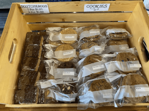 A wooden tray filled with packaged scotcheroos and various cookies, labeled with prices.