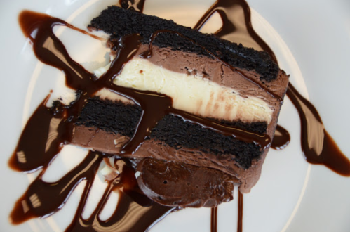 A slice of chocolate cake with layers of cream, drizzled with chocolate sauce on a white plate.