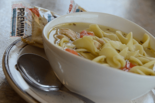 A bowl of chicken noodle soup with vegetables, accompanied by crackers on a plate and a spoon.