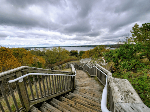 Wooden stairs lead down through trees, with a cloudy sky and river visible in the distance.