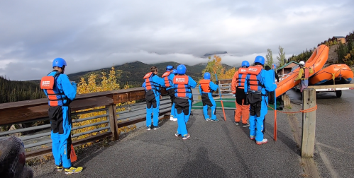 A group of people in blue and orange rafting gear prepares by a river, surrounded by autumn foliage and mountains.