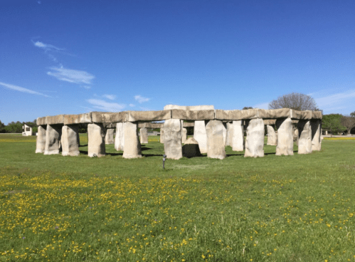 A replica of Stonehenge made of large stone blocks, set in a grassy field under a clear blue sky.