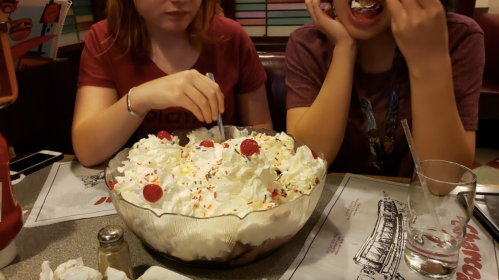 Two people enjoy a large ice cream sundae topped with whipped cream, cherries, and sprinkles at a diner.