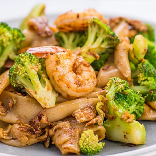 A plate of stir-fried noodles with shrimp and broccoli, garnished with green vegetables.