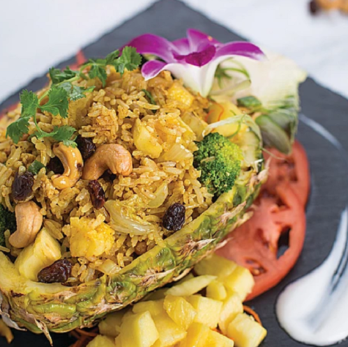A vibrant dish of pineapple fried rice served in a pineapple shell, garnished with herbs, nuts, and colorful vegetables.
