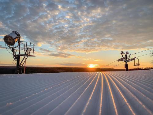 A sunset view over a snow-covered slope, with ski lift towers and clouds in the sky.
