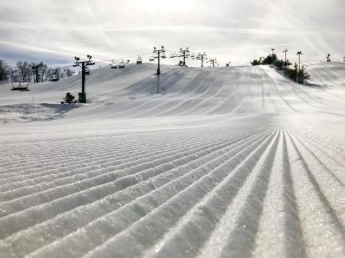 A snowy ski slope with groomed lines, ski lifts in the background under a bright sky.