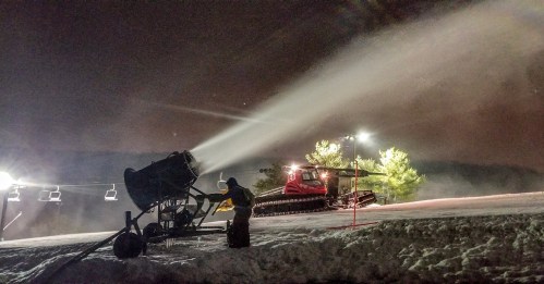 A snowmaking machine operates at night, with a snowcat in the background and ski lift lights illuminating the scene.