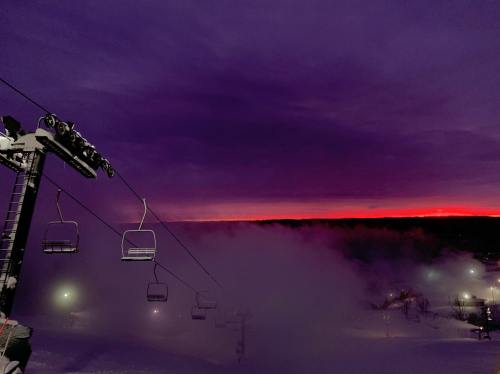 A ski lift silhouetted against a purple and orange sunset, with fog rolling over a snowy landscape.