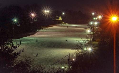 A nighttime view of a lit ski slope with skiers gliding down, surrounded by trees and glowing lights.