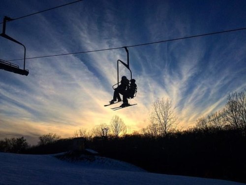 A silhouetted skier on a chairlift against a colorful sunset sky with wispy clouds.