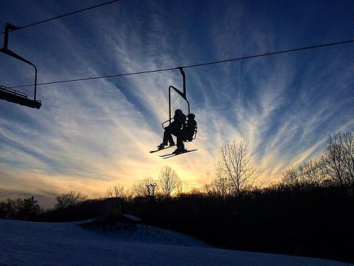 A silhouette of a skier on a chairlift against a colorful sunset sky with wispy clouds.