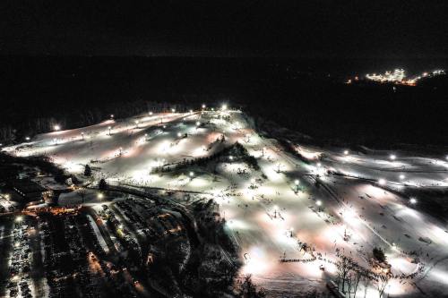 Aerial view of a lit ski resort at night, with skiers on slopes and a parking lot below.