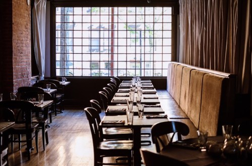 A cozy restaurant interior with wooden tables, chairs, and large windows letting in natural light.