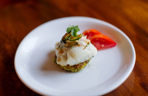 A plated dish featuring a fish fillet on a bed of green crust, garnished with herbs and served with tomato slices.